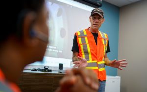 man in high-visibility vest and baseball cap presenting in front of a large screen